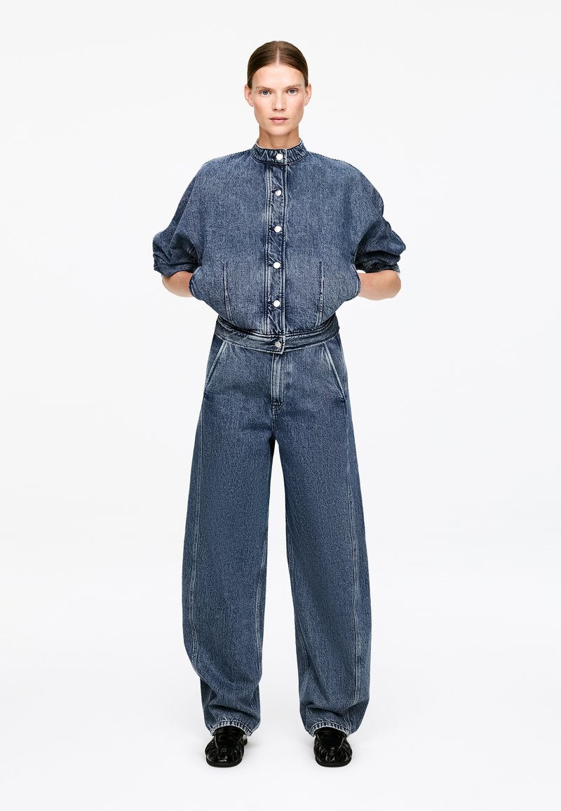 Femme debout portant une veste en denim bleu foncé ample avec des manches bouffantes et un jean large assorti, mains dans les poches, chaussures noires.