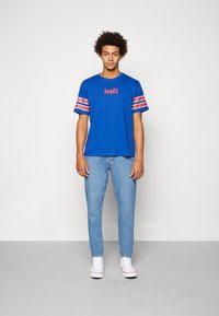 Camiseta de algodón azul con el logo rojo de "Levi's", con mangas a rayas blancas y rojas. Combinada con jeans azul claro y zapatillas blancas.