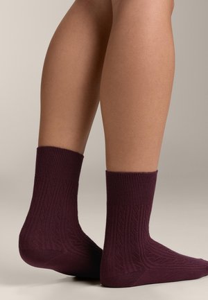Legs wearing mid-calf, textured dark purple socks standing on a neutral background with one foot slightly raised.