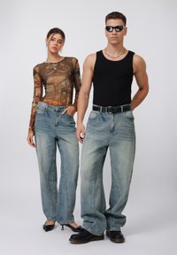 Dos modelos posan con jeans de mezclilla azul claro de corte holgado. Uno lleva una camiseta de manga larga impresa, el otro una camiseta sin mangas negra y gafas de sol.