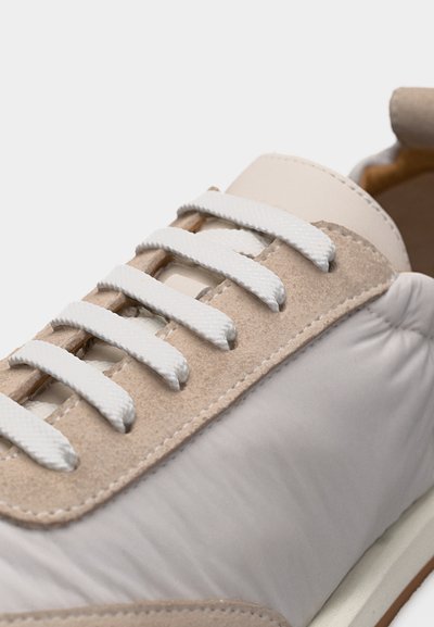 Κοντινό πλάνο μπεζ και λευκού αθλητικού παπουτσιού με λευκά κορδόνια και σουέντ λεπτομέρειες σε ανοιχτό φόντο.