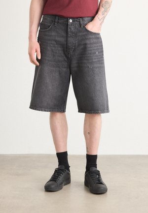 Homme portant un short en jean noir délavé, des chaussettes noires, des baskets noires et une chemise rouge avec une main dans la poche et des tatouages visibles sur les bras et les jambes.