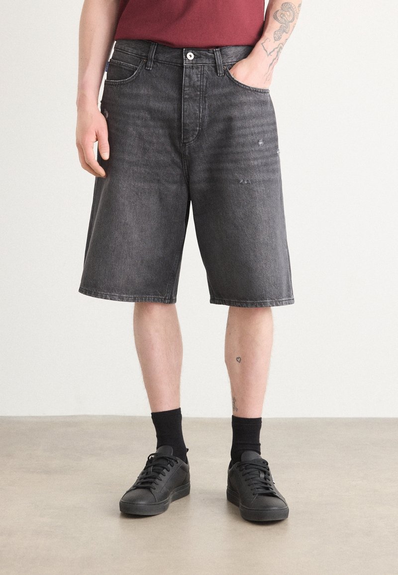Homme portant un short en jean noir délavé, des chaussettes noires, des baskets noires et une chemise rouge avec une main dans la poche et des tatouages visibles sur les bras et les jambes.