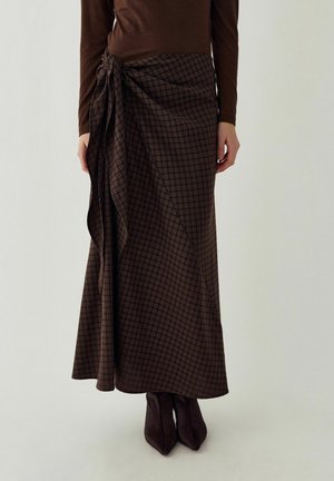 Jupe longue marron à carreaux nouée à la taille portée avec un haut marron à manches longues assorti et des bottes pointues sur fond blanc.
