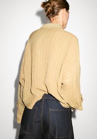 Camicia leggera, con texture, di colore senape, dal fit morbido e maniche lunghe, indossata sopra pantaloni in denim blu scuro con cuciture in vista.