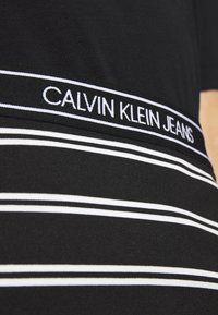 Svart bomull T-shirt med vita horisontella ränder och en svart midjeband med "CALVIN KLEIN JEANS" i vit text. Slät textur.
