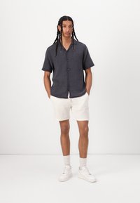Chemise à manches courtes, boutonnée, en tissu texturé gris foncé, accompagnée d'un short crème. Des baskets blanches décontractées complètent le look.