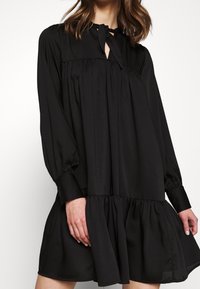 Vestido negro de manga larga con collar alto, corsé plisado y un dobladillo acampanado con volantes; tejido suave y silueta relajada.