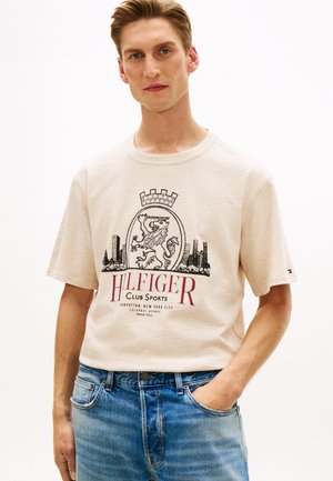 Man draagt beige T-shirt met korte mouwen met leeuwembleem en tekst "Hilfiger Club Sports", gecombineerd met blauwe jeans, hand in de zak.