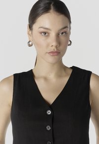 Jonge vrouw met donker haar in een lage paardenstaart draagt een zwart mouwloos topje met knopen en zilveren oorringen, en kijkt naar voren.