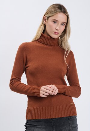 Lyshåret kvinde iført en rustfarvet rullekravesweater og sorte jeans, hænder løst sammenfoldet foran mod en ensfarvet baggrund.