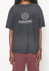 Černé oversized tričko vyrobené z měkké tkaniny, s centrální grafikou a textem "OCEANIC EXPLORATION" a emblémem koruny.