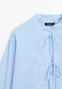 Blouse rayée verticalement bleu clair et blanc avec un col rond et deux nœuds noués sur le devant.