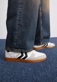 White sneakers with black stripes and a gum sole, featuring a mix of smooth leather and textured materials. Paired with denim jeans.