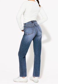 Džínové kalhoty s vysokým pasem v modré barvě, rovného střihu, s prominentními zadními kapsami a viditelným švem, v kombinaci s bílým tílkem s dlouhým rukávem.