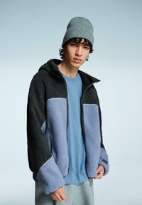 Fleecejack heeft een zwarte, grijze en lichtblauwe kleur met een rits aan de voorkant en een capuchon. Het wordt gecombineerd met een blauwe shirt en grijze broek. Zacht, gestructureerd materiaal.