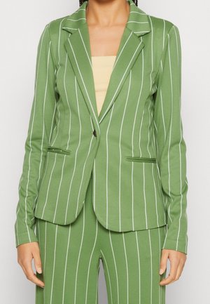 Femme vêtue d'un costume à rayures vertes avec un blazer à un bouton sur un top beige, debout devant un fond blanc uni.