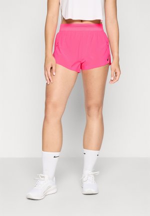 Femme portant un short de sport rose vif, des chaussettes blanches Nike, des sneakers blanches et un haut court blanc, le tout sur un fond clair.