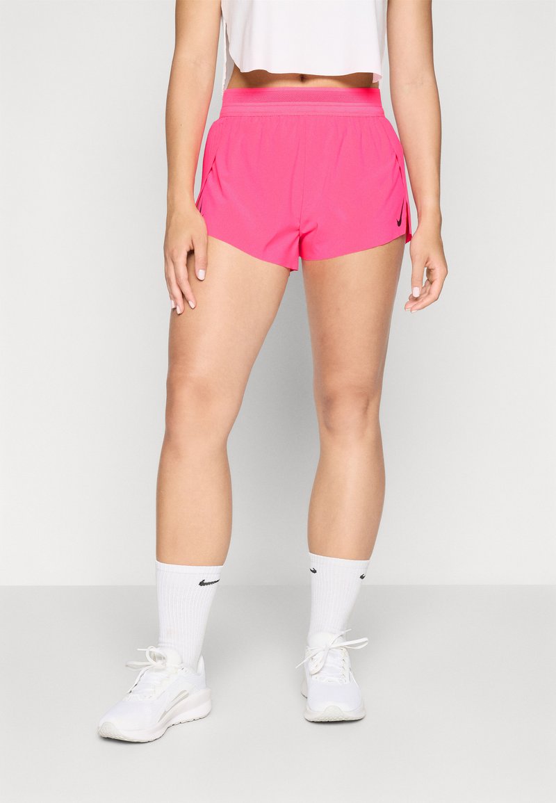 Eine Frau trägt leuchtend pinke Sportshorts, weiße Nike-Socken, weiße Turnschuhe und ein kurzes weißes Oberteil vor einem schlichten hellen Hintergrund.