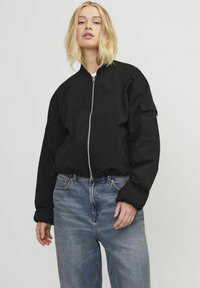 Svart bomberjakke med en strukturert design, glidelås og ribbede mansjetter. Båret over løstsittende blå jeans med en falmet tekstur.