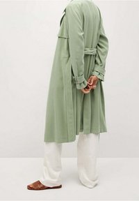 Jasnozielony płaszcz typu trench z wcięciem w talii i podwiniętymi mankietami, połączony z białymi spodniami o szerokich nogawkach i brązowymi klapkami.