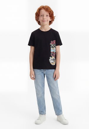 Garçon aux cheveux roux portant un t-shirt noir avec un motif graphique, un jean bleu clair et des baskets blanches, debout devant un fond blanc.