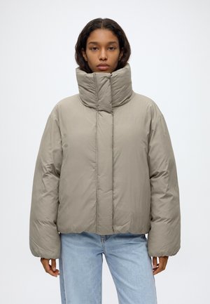 Mujer que lleva una chaqueta acolchada de cuello alto de color gris claro con mangas largas y vaqueros azul claro sobre un fondo blanco liso.