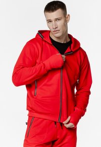 Czerwona bluza z kapturem na zamek błyskawiczny z czarnym wnętrzem i kieszeniami na zamek. Posiada długie rękawy z otworami na kciuki oraz logo na kołnierzu kaptura.