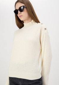 Maglione a maglia color crema con colletto alto, dotato di tre bottoni dorati sulla spalla sinistra e bordo a coste. Tessuto morbido.