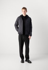 Casaco acolchoado preto com capuz, apresentando uma mistura de texturas lisas e acolchoadas, usado com calças e sapatos pretos. Design retangular simples.