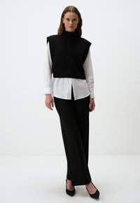Zwarte gebreide mouwloze top met hoge hals, gelaagd over een witte blouse met knopen, gecombineerd met zwarte slim-fit pants en spitse zwarte hakken.