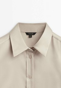 Chemise beige à boutons avec col pointu, manches longues et étiquette de marque noire visible à l'intérieur de l'encolure.