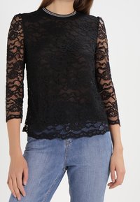 Top in pizzo nero con maniche a tre quarti e orlo scalloped. Presenta un colletto rotondo con rifinitura argentata a contrasto.