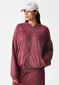 Bordo zip-up kapica s kapuco, velikim vezenjem "NEW ERA", sprednjimi žepi in rebrastimi manšetami. V kombinaciji z ujemajočo se krilom.