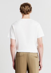 Top corto in maglia bianca a maniche corte con scollo rotondo, abbinato a pantaloni di un marrone chiaro con tasche laterali e una texture liscia.