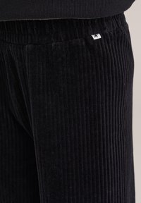 Čierne kordové nohavice majú vertikálne pruhy, elastický pás a malú značkovú nálepku na boku na identifikáciu.