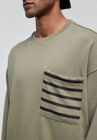 Olijfgroene sweatshirt met een ronde halslijn en een borstzak aan de linkerkant met zwarte horizontale strepen, gemaakt van zachte, gestructureerde stof.