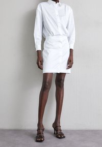 Una persona è in piedi contro un muro semplice indossando un vestito camicia bianco con bottoni e sandali con tacco alto neri su un pavimento grigio.