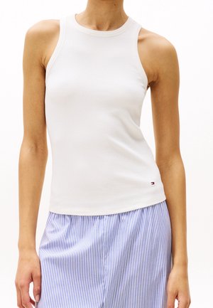 Femme portant un haut blanc sans manches côtelé associé à un pantalon rayé verticalement bleu et blanc sur un fond uni.