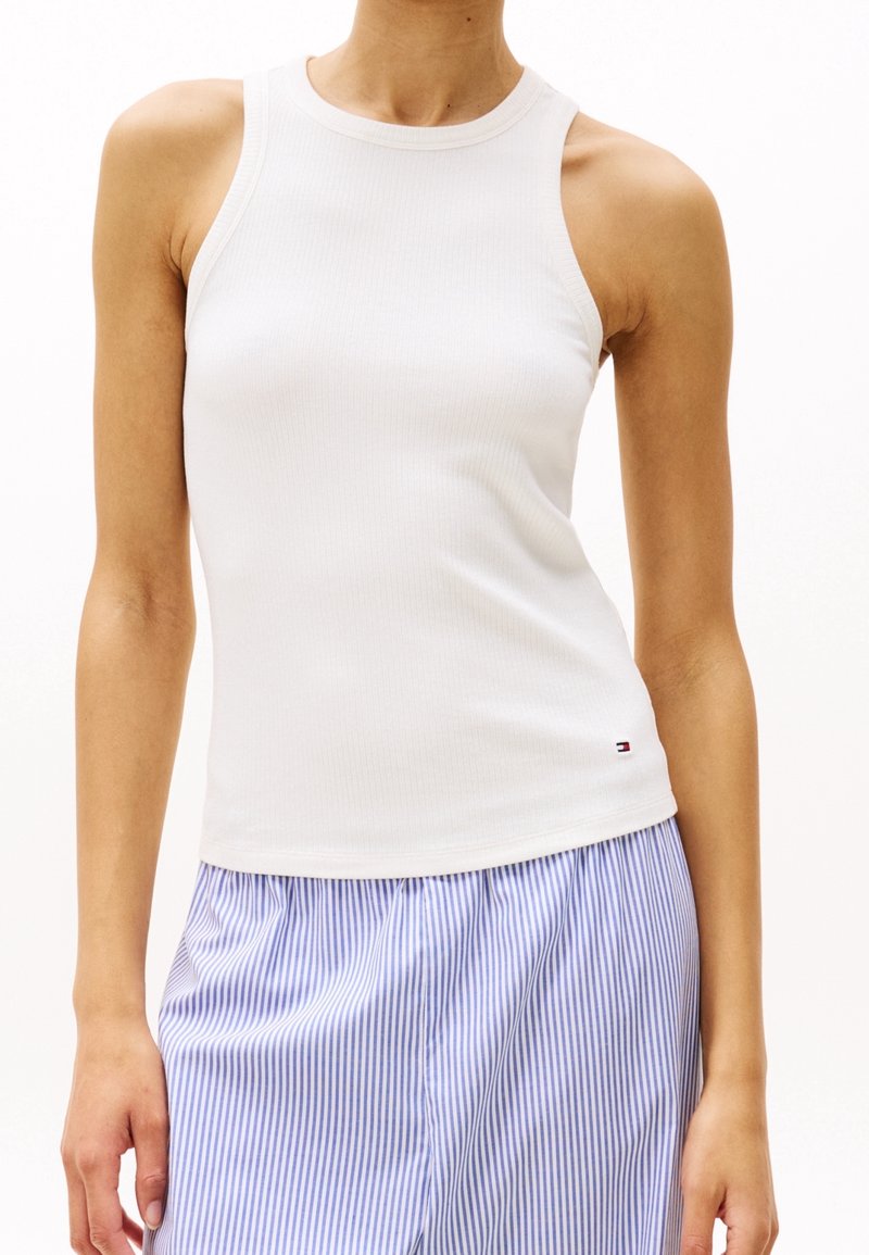 Femme portant un haut blanc sans manches côtelé associé à un pantalon rayé verticalement bleu et blanc sur un fond uni.