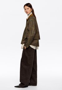 Kurtka w oliwkową kratę z warstwowym designem i zapinanymi mankietami, zestawiona z luźnymi brązowymi spodniami oraz jasnym golfem pod spodem.