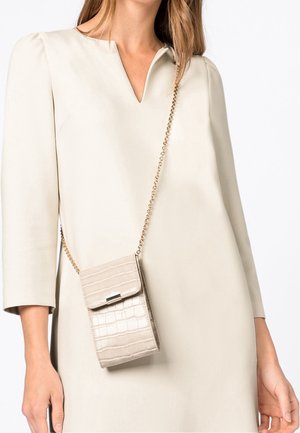 Femme portant une robe beige à col en V avec des manches trois-quarts, portant un petit sac bandoulière beige à texture crocodile avec une sangle chaîne dorée.