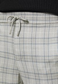 Lichtgrijze pluche joggers met blauwe accenten, een tailleband met trekkoord en een metalen punt, met een gestructureerd stoffenpatroon.