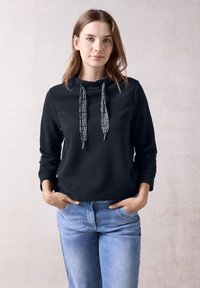 Svart huvtröja med strukturerad tyg, med mönstrad dragsko och sidofickor, kombinerad med ljusblå jeans.