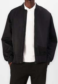 Veste bomber noire avec fermeture éclair à l'avant, poignets et col côtelés. Texture lisse, tissu léger, associée à un polo blanc en dessous.