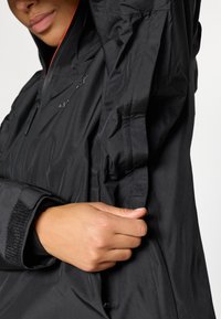 Schwarze, wetterfeste Jacke mit einem Reißverschluss vorne, verstellbaren Bündchen mit Klettverschluss und einem schlanken Design mit Kapuze.