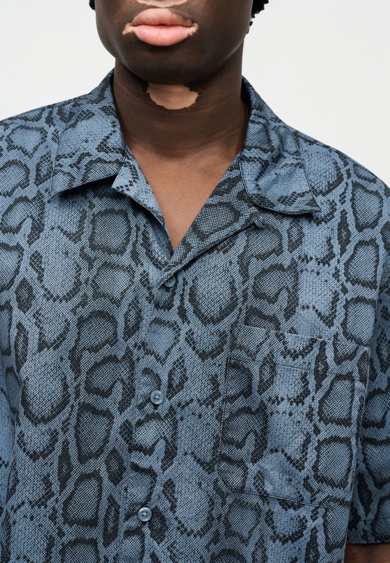 Camisa de manga corta con estampado de serpiente en azul y negro, que presenta un cierre de botones en el frente, un cuello y un bolsillo en el pecho con una textura suave.