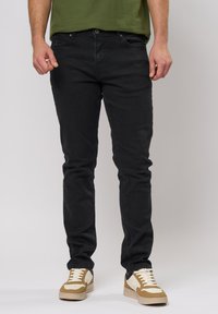 Jean en denim noir avec une coupe slim, présentant des poches avant, une fermeture à bouton et un tissu texturé subtil. Associés à des baskets beige et crème.