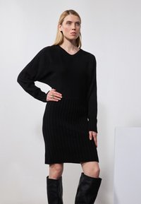 Schwarzes Strickkleid mit V-Ausschnitt, gerippter Textur, langen Ärmeln und figurbetonter Silhouette. Das Model trägt hohe schwarze Stiefel.