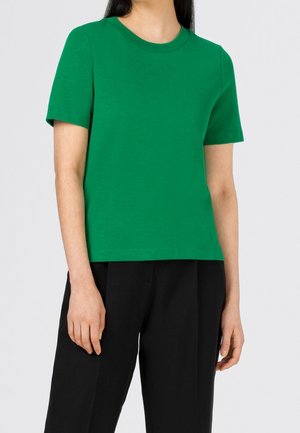 Frau trägt ein schlichtes grünes Kurzarmshirt und lockere schwarze Hose, steht vor einem neutralen hellen Hintergrund.